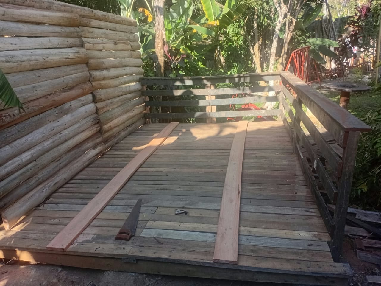 Deck de madeira rústica em fase final de construção na chácara, com parede de toras e jardim tropical ao fundo.