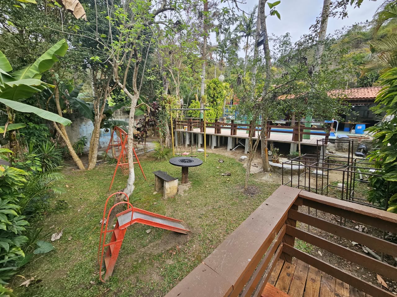 Área de lazer da chácara com playground infantil, escorregador, balanços, deck de madeira e casa ao fundo em meio a muito verde.