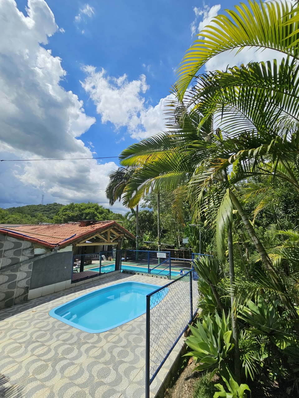 Área da piscina com quiosque coberto, deck azulejado e vegetação tropical, ideal para mostrar a área de lazer da chácara.
