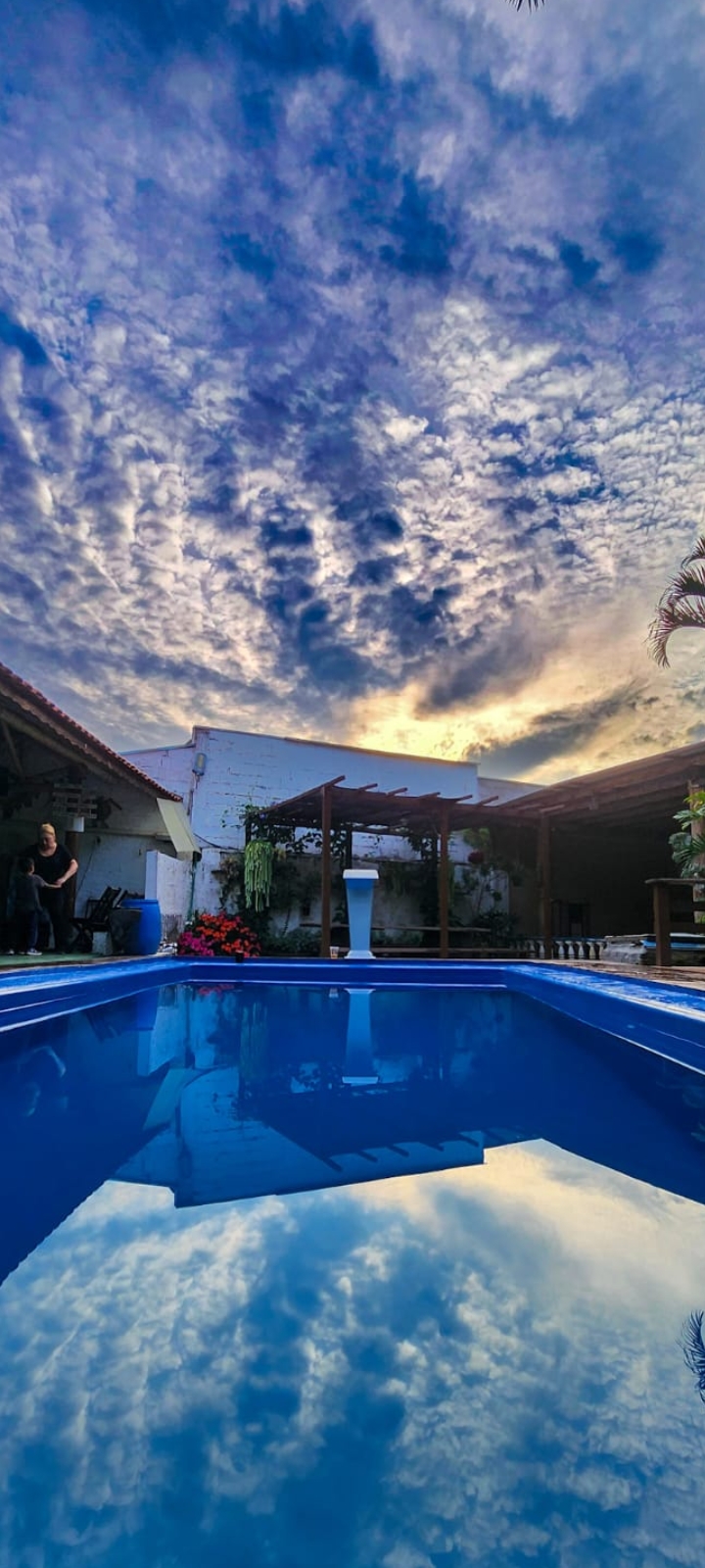 Piscina da chácara ao entardecer, com pergolado, plantas e céu dramático, mostrando a área de lazer para hospedagem e eventos.