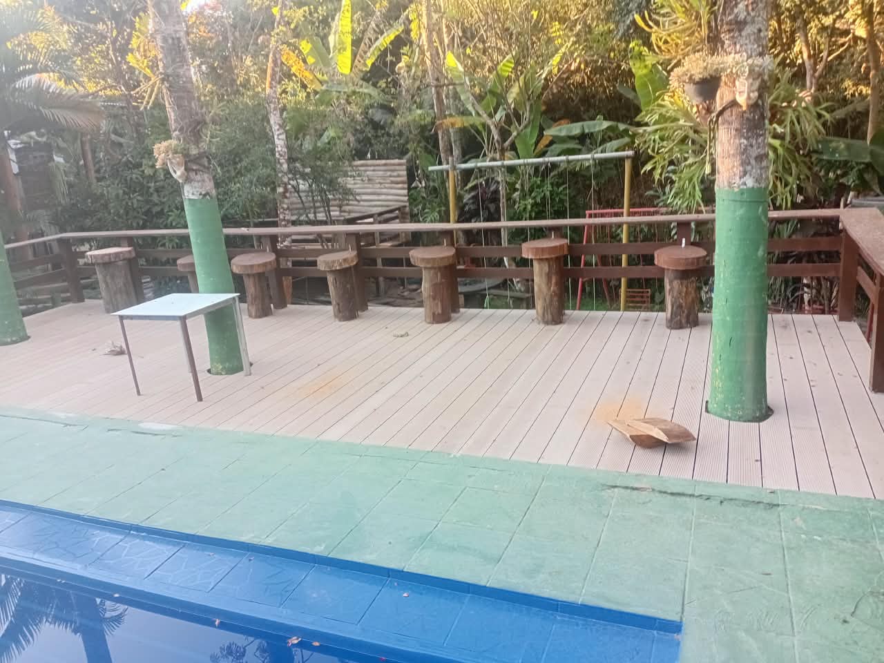 Deck de madeira com banquetas rústicas, mesa, piscina e área de lazer integrada à mata tropical da chácara.