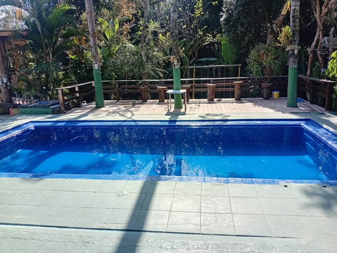 Piscina ampla com deck de madeira e área de descanso rodeada por vegetação tropical, uma das áreas de lazer da chácara.