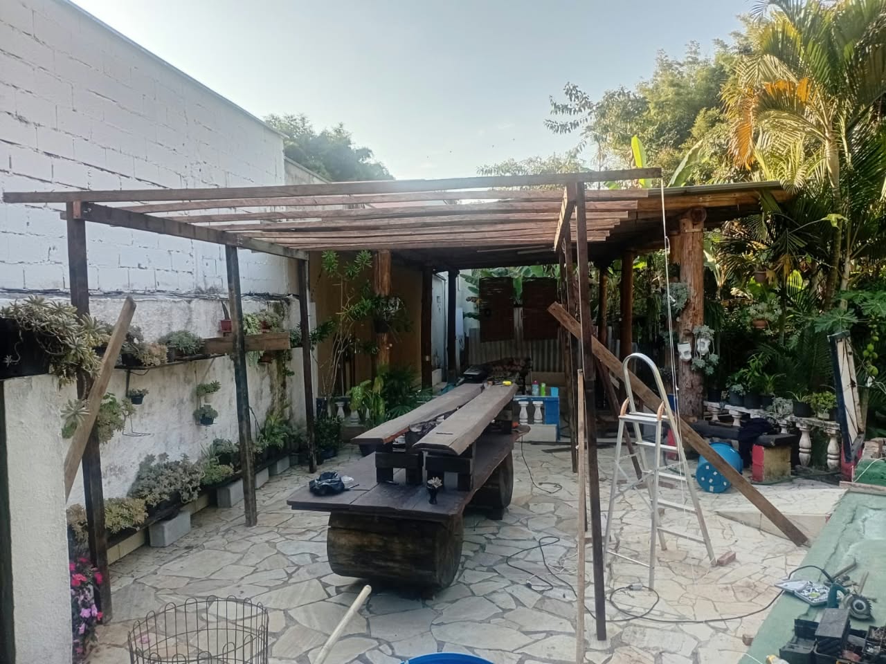 Área de lazer externa da chácara com pergolado de madeira, mesa rústica, plantas decorativas e piscina ao fundo.